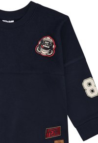 Felpa blu navy con texture e patch di un astronauta con la scritta "OUT of this WORLD", numero "8" bianco sulla manica e patch rossa "YEAH!" vicino all'orlo.