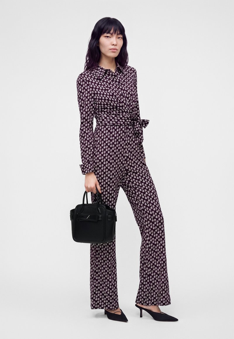 Femme portant une combinaison noire et blanche à motif avec une ceinture à la taille, tenant un sac à main noir et portant des talons pointus noirs.