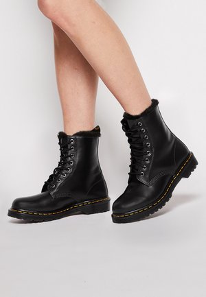 Jambes portant des bottines en cuir noir à lacets, doublées de fourrure et dotées de surpiqûres jaunes sur des semelles épaisses, sur un fond clair uni.
