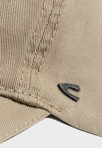 Beige corduroy kasket med struktureret stof, syede detaljer og en sort logoaccents på siden.