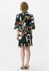 Robe courte, noire, à motifs avec des manches bouffantes et une taille nouée. Présente des couleurs abstraites en jaune, bleu et blanc. Texture de tissu lisse.