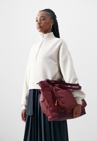 Bordeaux platnena torba z urejenim oblikom, nastavljivimi naramnicami in sprednjim žepom. Nosijo jo oseba v kremastem puloverju in črni zloženi krili.