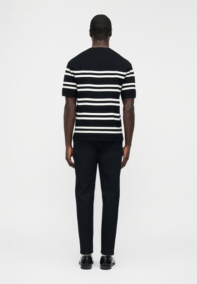 Άνδρας που στέκεται με την πλάτη προς τα εμπρός, φορώντας μαύρο και λευκό κοντομάνικο πουκάμισο με οριζόντιες ρίγες, μαύρο παντελόνι και μαύρα παπούτσια σε ουδέτερο φόντο.