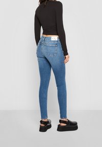Mujer vestida con pantalones ajustados de talle alto azul, blusa negra de manga larga y zapatos de plataforma negros, de pie contra un fondo blanco liso.