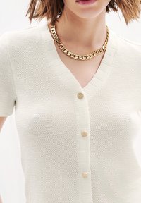 Cárdigan de punto crema con escote en V, mangas cortas y tres botones dorados texturizados en la parte delantera. Un collar de cadena dorada añade un toque de acento.