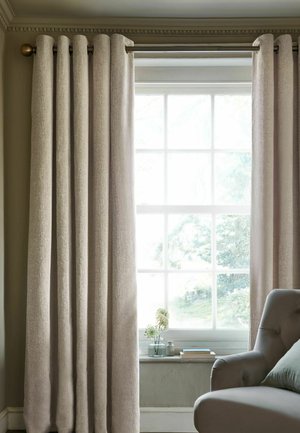 Rideaux texturés beiges partiellement ouverts sur une fenêtre avec des plantes, des livres sur le rebord et un fauteuil rembourré gris à proximité.