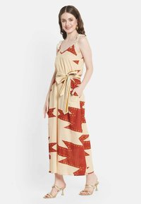 Robe maxi beige avec un motif zigzag rouge, dotée de bretelles volantes, d'une taille à nouer et de poches latérales. Tissu lisse avec une silhouette fluide.