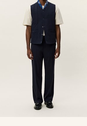 Homme portant une veste sans manches bleu marine sur une chemise beige à manches courtes, portée avec un pantalon bleu marine et des chaussures noires épaisses.