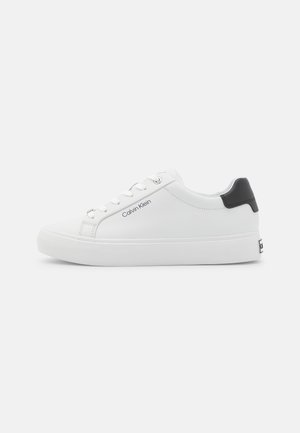 Zapatilla blanca con parte superior de cuero liso, puntera redondeada, acento negro en el talón y logo en el lateral. Suela gruesa de goma blanca, diseño minimalista.