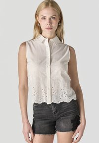 Blonde vrouw die een mouwloze witte geborduurde blouse met knopen draagt en zwarte denim shorts met bloemrijke kanten details op de dijen.