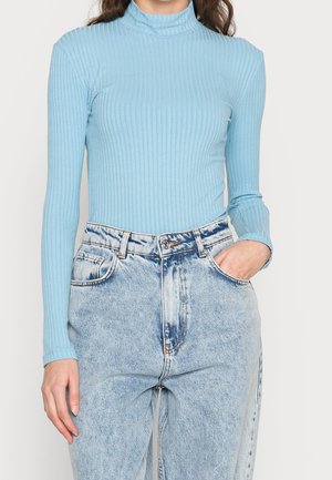 Frau trägt einen hellblauen gerippten Langarm-Rollkragenpullover, in hoch taillierte Acid-Wash-Jeans gesteckt, mit einer Hand in der Tasche.