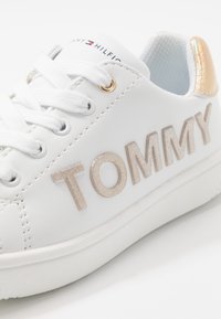 Zapatillas sintéticas blancas con acentos dorados y la palabra "TOMMY" en letras metálicas brillantes. Presenta un diseño con cordones y suela texturizada.