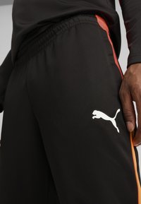 Calças desportivas pretas com um cós elástico, apresentando riscas laterais laranjas e um logótipo branco da Puma na coxa esquerda. Textura de tecido suave.