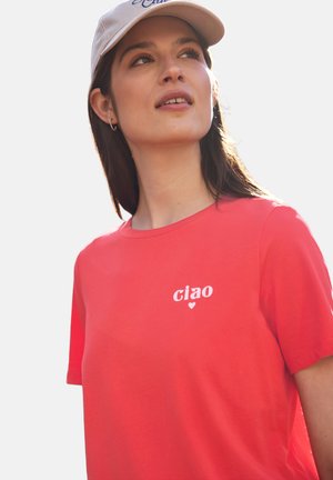 Femme portant un T-shirt rouge avec « ciao » et un petit cœur imprimé, regardant vers le haut, avec de longs cheveux foncés et une casquette beige.