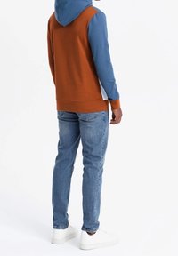 Sudadera con bloques de color en paneles de óxido y azul, con capucha, puños acanalados y un bolsillo tipo canguro. Combinada con jeans azul claro y zapatillas blancas.