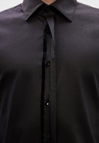 Fekete ing, sima textúrával, fényes felülettel, bársonyos gombolópánttal és fekete gombokkal. Vékony gallér és testhezálló kialakítás.
