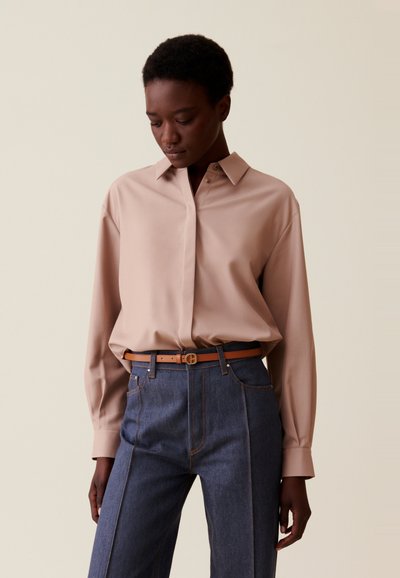 Persona con una camisa de manga larga beige metida dentro de unos pantalones vaqueros de cintura alta de color azul oscuro, con un cinturón marrón claro, mirando hacia abajo.