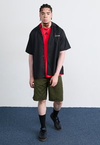 Black short-sleeve shirt over a red t-shirt, olive green shorts, black shoes with laces, and black socks. Simple design with no patterns.