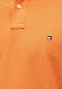 Πορτοκαλί πόλο μπλουζάκι από υφασμάτινη υφή. Διαθέτει τρία κουμπιά στη λαιμόκοψη και μικρό κεντητό λογότυπο σε κόκκινο, λευκό και μπλε.