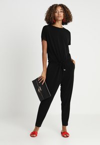 Zwarte jumpsuit met een knoopdetail op de taille, korte mouwen en zijzakken. Gecombineerd met rode sandalen en een zwarte clutch.