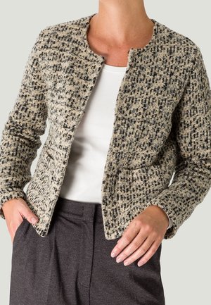 Personne portant une veste beige et noire texturée et à motifs sur un haut blanc avec un pantalon gris foncé, debout avec une main dans la poche.
