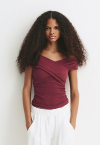 Jeune femme avec de longs cheveux bouclés portant un haut bordeaux à épaules dénudées et un pantalon blanc, se tenant devant un fond blanc avec les mains dans les poches.