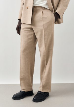 Beige op maat gemaakte broek gecombineerd met een bijpassend blazer en zwarte lederen loafers, gedragen door een staande persoon met handen in de zakken.