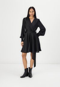 Robe noire en tissu lisse, dotée de manches longues, d'une taille cintrée avec un lien et d'une jupe évasée qui tombe au-dessus du genou.
