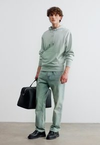 Νεαρός με κατσαρά μαλλιά που φοράει μια ανοιχτό γκρι μπλούζα με κουκούλα, ξεθωριασμένο μπλε τζιν, μαύρα παπούτσια και κρατά μια μαύρη ταξιδιωτική τσάντα στο αριστερό του χέρι.