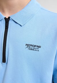 Ljusblå polo skjorta med dragkedja vid kragen, med svart text "PETROLIND Miami beach FLORIDA, U.S.A." på vänster sida.
