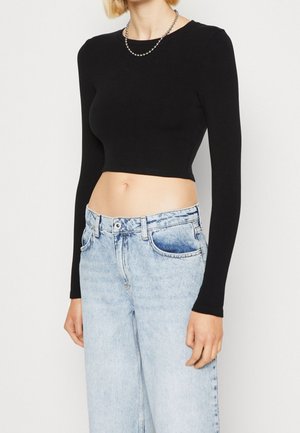 Kobieta w czarnym, długim rękawem, krótkim topie, jasno-niebieskich, wysokowtalowych jeansach i srebrnym naszyjniku z koralików, stoi na białym tle.