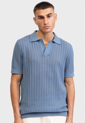 Άνδρας που φοράει ανοιχτό γαλάζιο πόλο με κοντά μανίκια και γιακά, καθώς και λευκό παντελόνι, στέκεται μπροστά σε απλό λευκό φόντο.