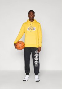 Yellow hoodie with "Los Angeles Lakers" logo, gray sweatpants featuring Lakers graphics, and white athletic shoes. Holding a basketball.