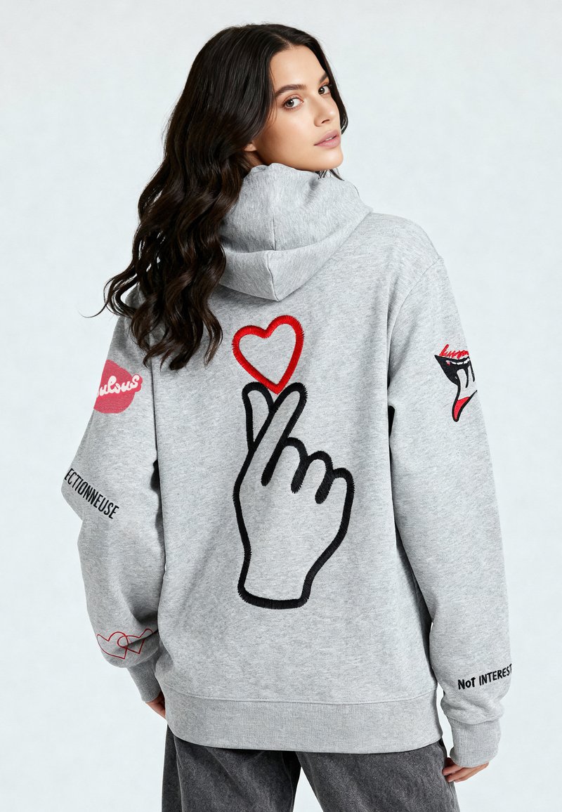 Hoodie gris avec un grand graphique de main avec un cœur au-dessus, détails brodés en rouge et noir, et texte sur les manches.