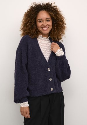 Navy fuzzy cardigan with a deep V-neck, four decorative buttons, and wide sleeves, layered over a white lace top with ruffled cuffs.