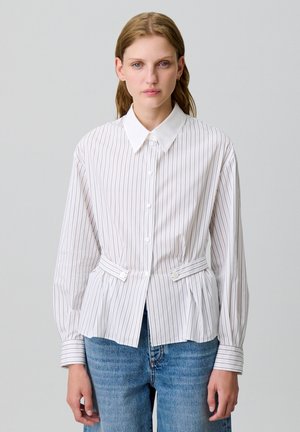 Chemise blanche à boutons avec des rayures verticales, coupe ajustée, col pointe et liens latéraux ; associée à un jean bleu clair.
