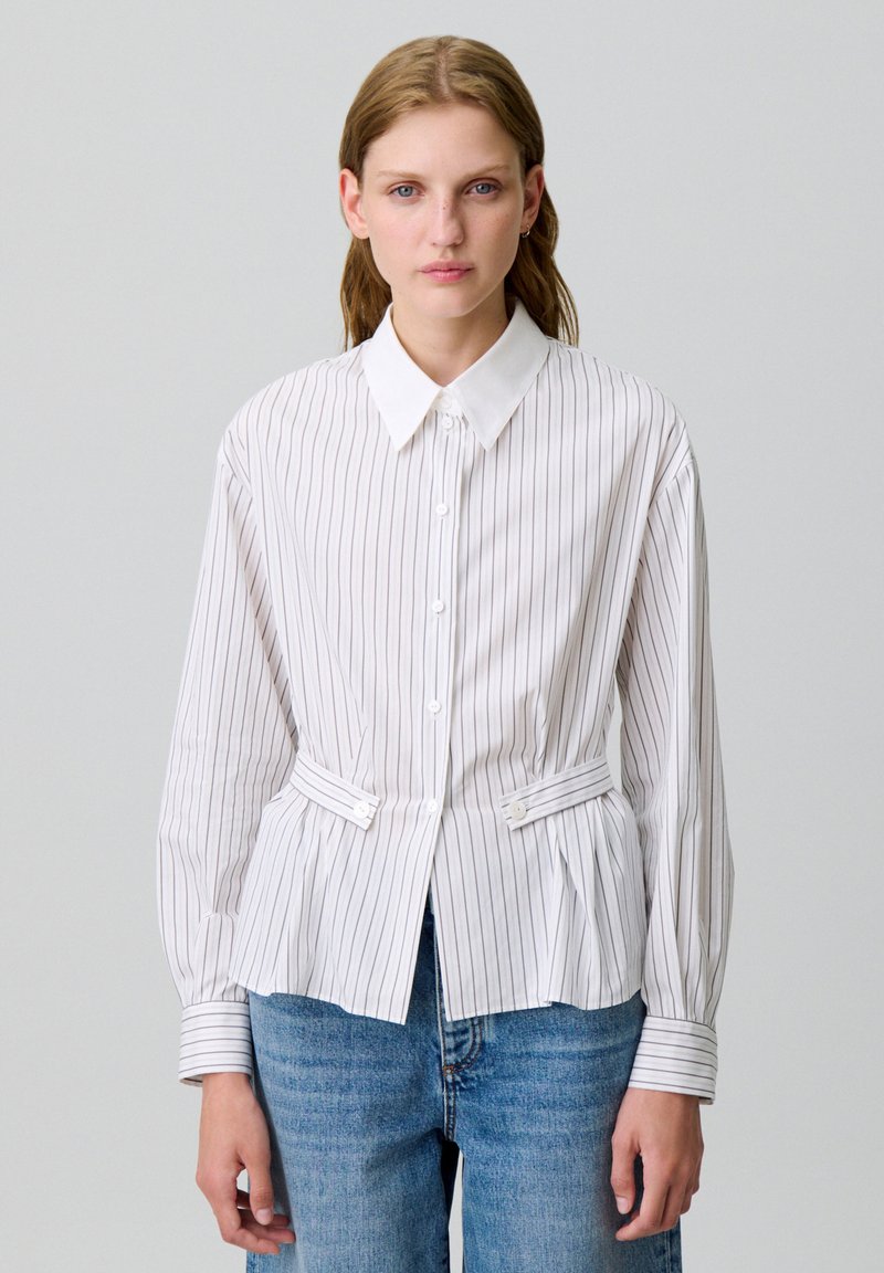 Camicia bianca con righe verticali, vestibilità slim, colletto a punta e lacci laterali; abbinata a jeans azzurri.