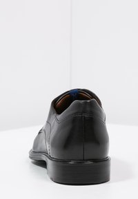 Scarpa da uomo in pelle nera con finitura liscia, punta arrotondata, cuciture minime e un piccolo accento blu sul tallone.