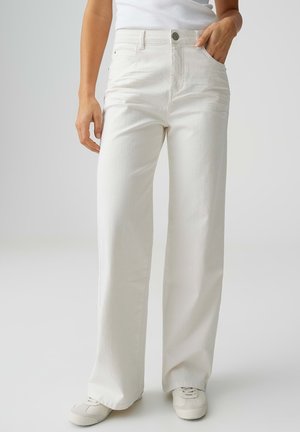 Person, die weiße Jeans mit geradem Bein, weiße Turnschuhe und ein weißes ärmelloses Oberteil trägt, mit einer Hand in der Tasche.