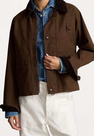 Persona che indossa una giacca marrone sopra una camicia di denim blu e pantaloni bianchi, mano che sistema la giacca vicino al petto.