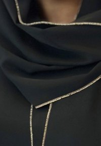 Mustaa kangasta, jossa on herkkä kultareunus reunoilla, lasketaan pehmeiden laskosten esittelemiseksi ja pieni osa ihoa kaula-aukon kohdalla.