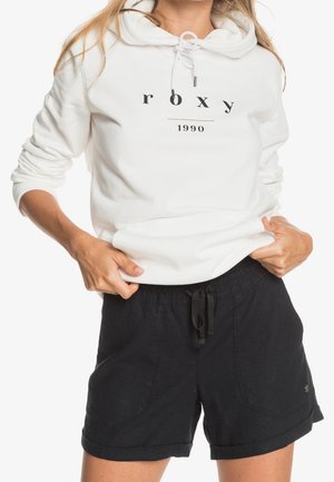 Kvinde iført en hvid hættetrøje med teksten "roxy 1990" og sorte snøre-shorts, hænder justerer hættetrøjens kant.