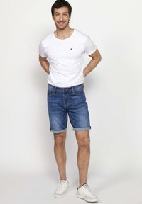 Camiseta blanca de manga corta con un pequeño logo en forma de triángulo, pantalones cortos de mezclilla azul con los dobladillos enrollados y zapatillas blancas. Un atuendo simple y casual.