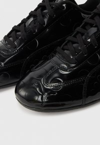 Schwarze Sneakers aus Lackleder mit erhabenen kreisförmigen Mustern und schwarzen Schnürsenkeln auf weißem Hintergrund, Nahaufnahme des Zehenbereichs.