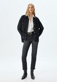 Chaqueta de mezclilla negra con dos bolsillos frontales, llevada sobre una camisa blanca, combinada con jeans de mezclilla negros y botas negras hasta el tobillo.