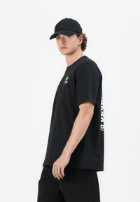 T-shirt en coton noir à manches courtes, avec un logo vert à l'avant et du texte blanc sur le côté. Associé à une casquette noire.