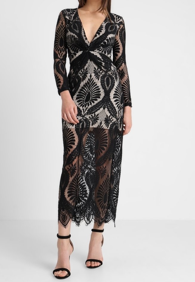 Femme portant une robe midi noire en dentelle à manches longues avec un profond décolleté en V et des motifs floraux complexes, associée à des talons noirs à bride à la cheville.