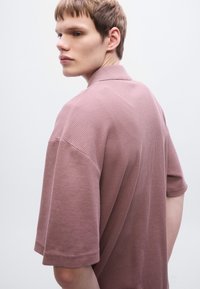 Πόλο μπλουζάκι με κοντό μανίκι σε υφασμα με υφή σε ροδακινί απόχρωση, διαθέτει γιακά και άνετη εφαρμογή. Η πλάτη παρουσιάζει μια διακριτική ραφή.