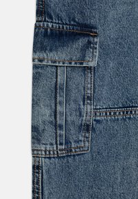 Denim tkanina v srednje modri barvi, ki vsebuje veliko naramno žep s vidnimi šivi in rahlo uničen teksturo.