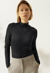 Frau mit kurzem blonden Haar, die goldene Creolen, einen schwarzen Langarmrollkragenpullover und beige Hosen trägt, mit den Händen in den Taschen vor einem hellen Hintergrund.